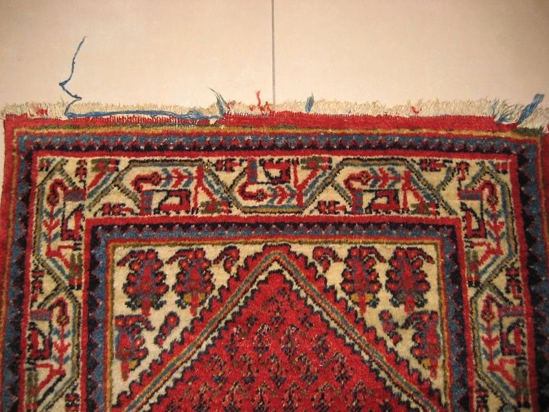 Damaged rug showing missing and worn fringes, awaiting restoration.